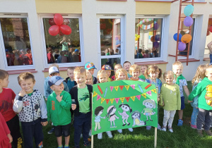 Dzieci stoją przed wejściem do przedszkola, prezentują plakat i śpiewają „Hymn Przedszkolaka”.
