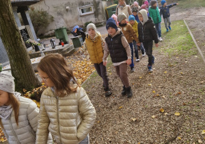 Przedszkolaki maszerują alejkami cmentarza przyglądając się grobom.