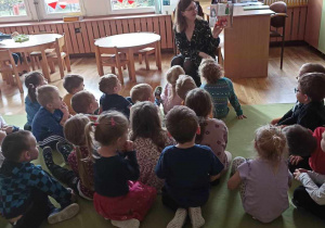 Dzieci siedzą na dywanie i słuchają wierszy Juliana Tuwima czytanych przez nauczyciela.